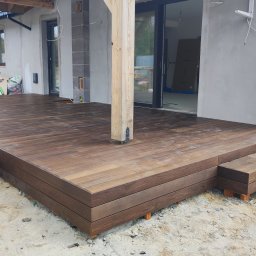 WIDAM Adam Wicha Usługi Remontowo-Budowlane - Nowoczesny taras z drewna kompozytowego w odcieniach brązu, z niskimi schodkami i elementami konstrukcyjnymi dachu, na tle surowego budynku w trakcie wykończenia.