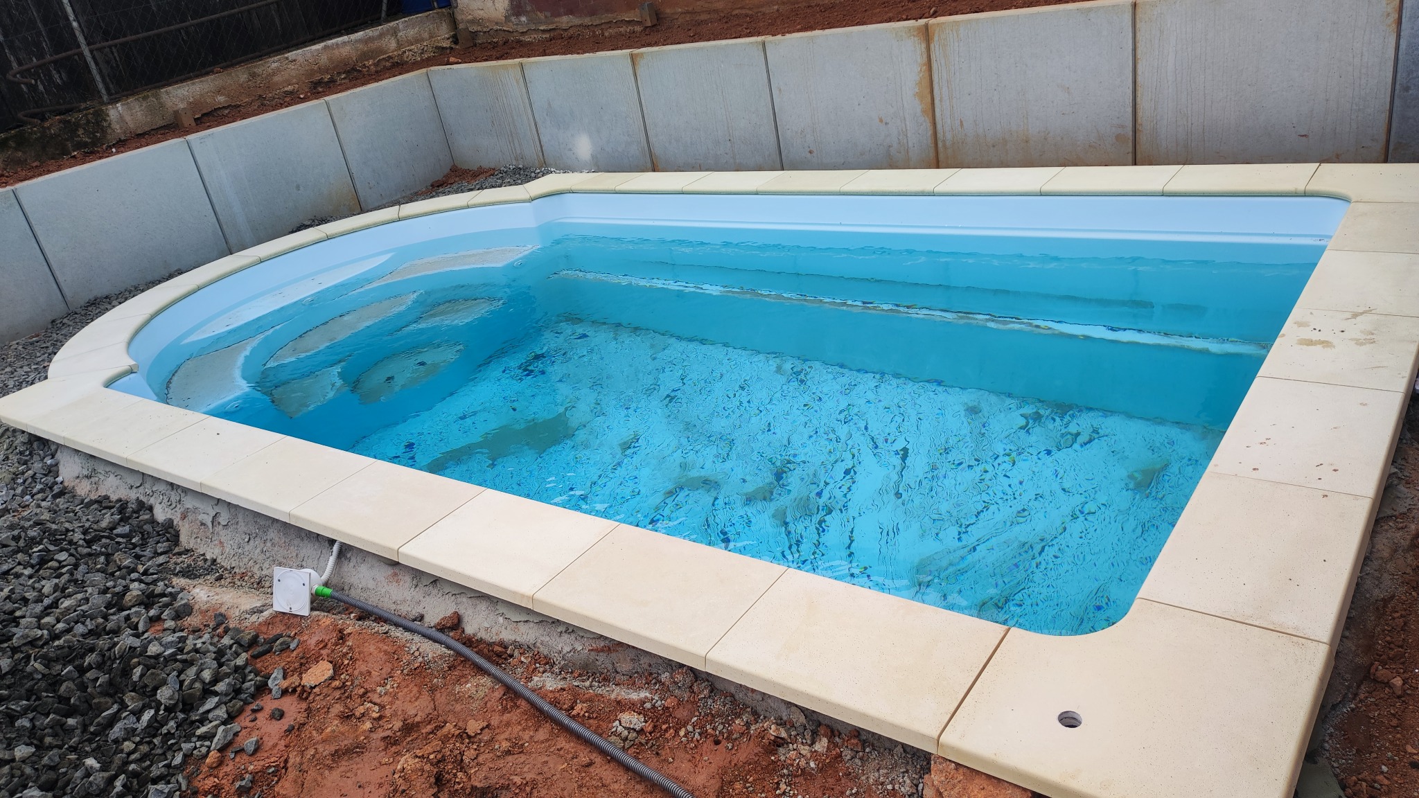 Nowoczesny basen z jasną mozaiką, wykończony kremowymi płytami, otoczony ziemią i kamieniami, z widocznym przyłączem elektrycznym.