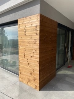 Drewniana obudowa narożna na zewnątrz budynku, z widocznym fragmentem okna i przesuwnych drzwi balkonowych, oraz częścią tarasu. Widoczne poziome deski z naturalnym usłojeniem drewna.