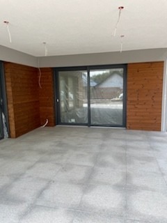 Wnętrze w trakcie prac wykończeniowych: szara, matowa podłoga z płytek, drewniana boazeria na ścianach, przesuwne okno tarasowe z ciemną ramą, wystające kable z sufitu.