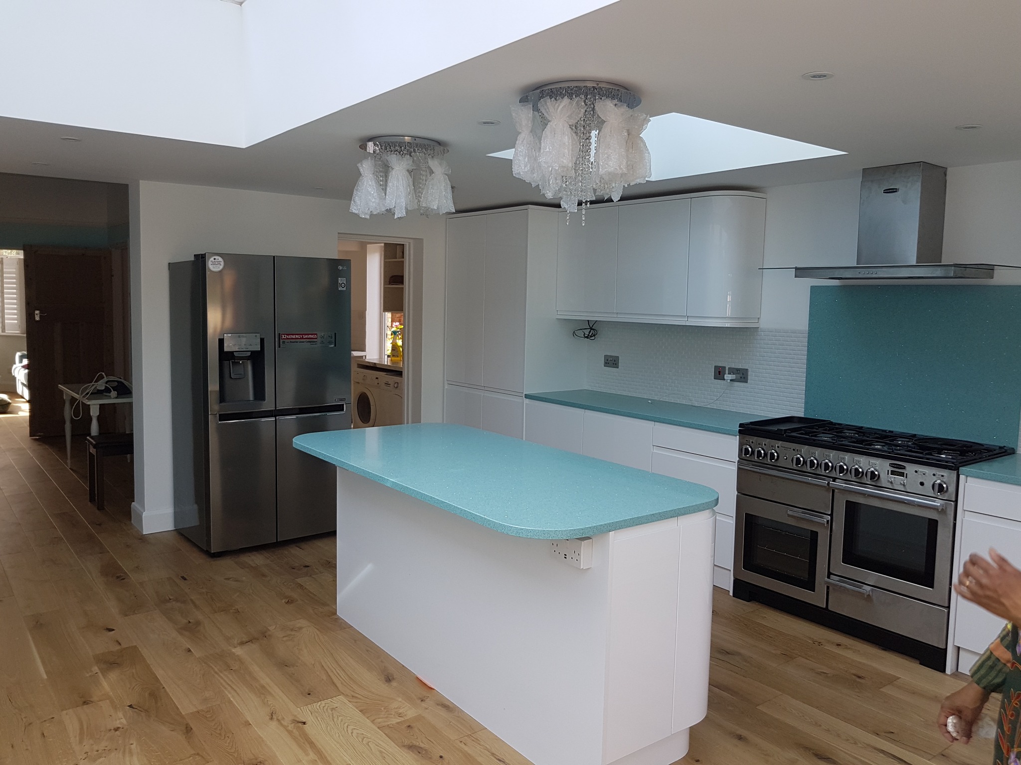 Jasna, przestronna kuchnia z wyspą o turkusowym blacie, białe szafki z zaokrąglonymi krawędziami, duża lodówka side-by-side, kuchenka z piekarnikiem i okapem, drewniana podłoga, lampy w folii...