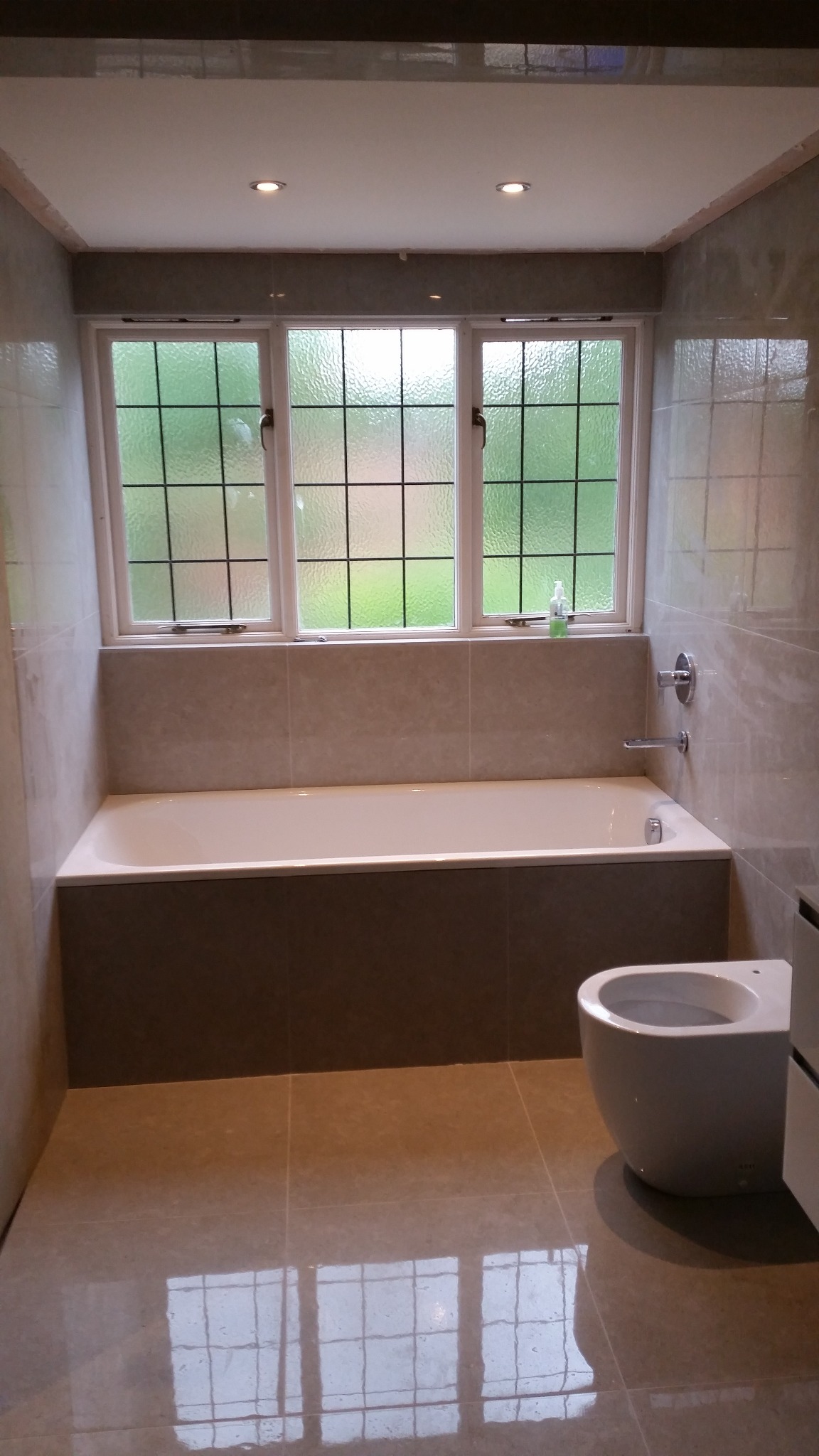 Wykończona łazienka z wanną obudowaną płytkami, wiszącą toaletą i oknem z kratownicą, wykończona beżowymi płytkami podłogowymi i ściennymi.