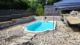 Niebieski basen ogrodowy w kształcie nerki, otoczony szarą kostką brukową i schodami z płyt kamiennych, z widocznymi elementami konstrukcyjnymi i zielenią w tle.