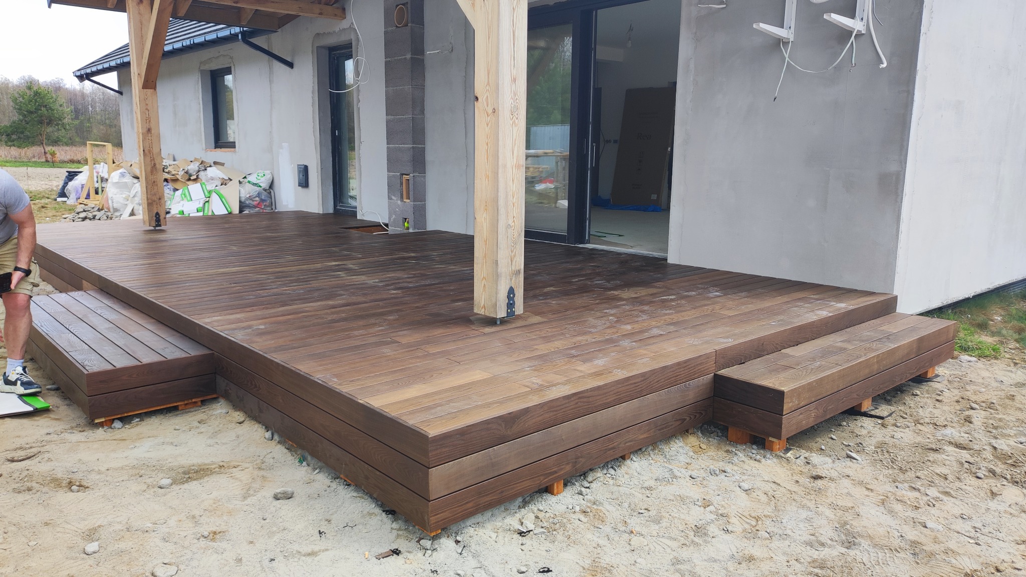 Nowoczesny taras z drewna kompozytowego w odcieniach brązu, z niskimi schodkami i elementami konstrukcyjnymi dachu, na tle surowego budynku w trakcie wykończenia.