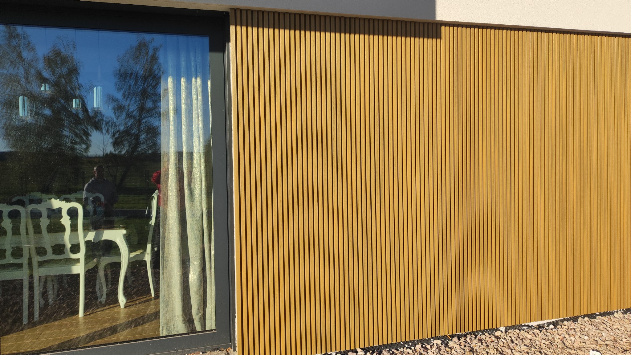 Nowoczesna elewacja domu z pionowych drewnianych paneli, przylegająca do okna tarasowego z widocznym odbiciem wnętrza: białe krzesła i stół w stylu Ludwika.