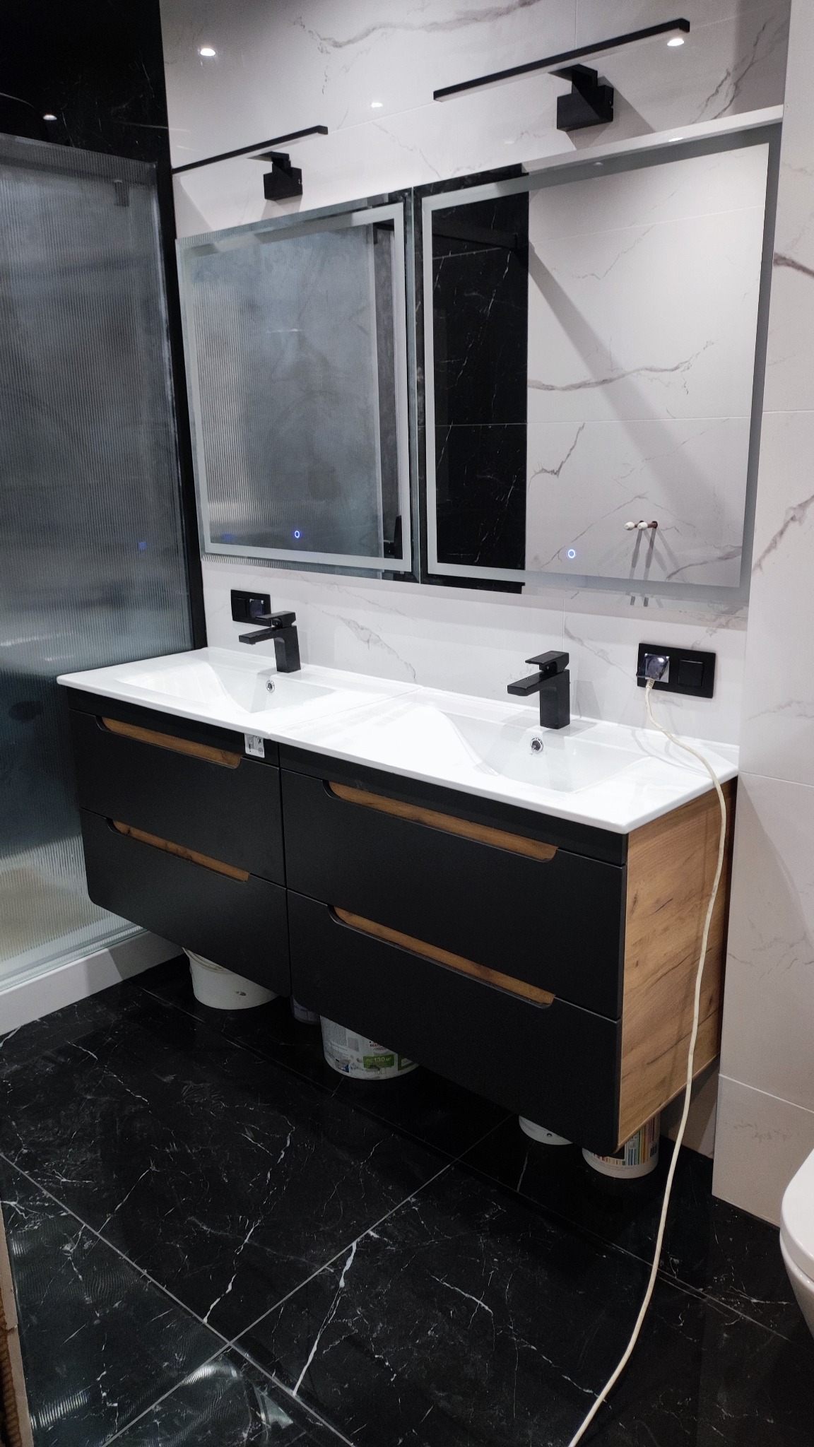 Nowoczesna łazienka z podwójną umywalką na szafce w kolorze czarnym i drewna, czarne baterie, lustra z podświetleniem LED i marmurowe płytki na ścianach i podłodze.