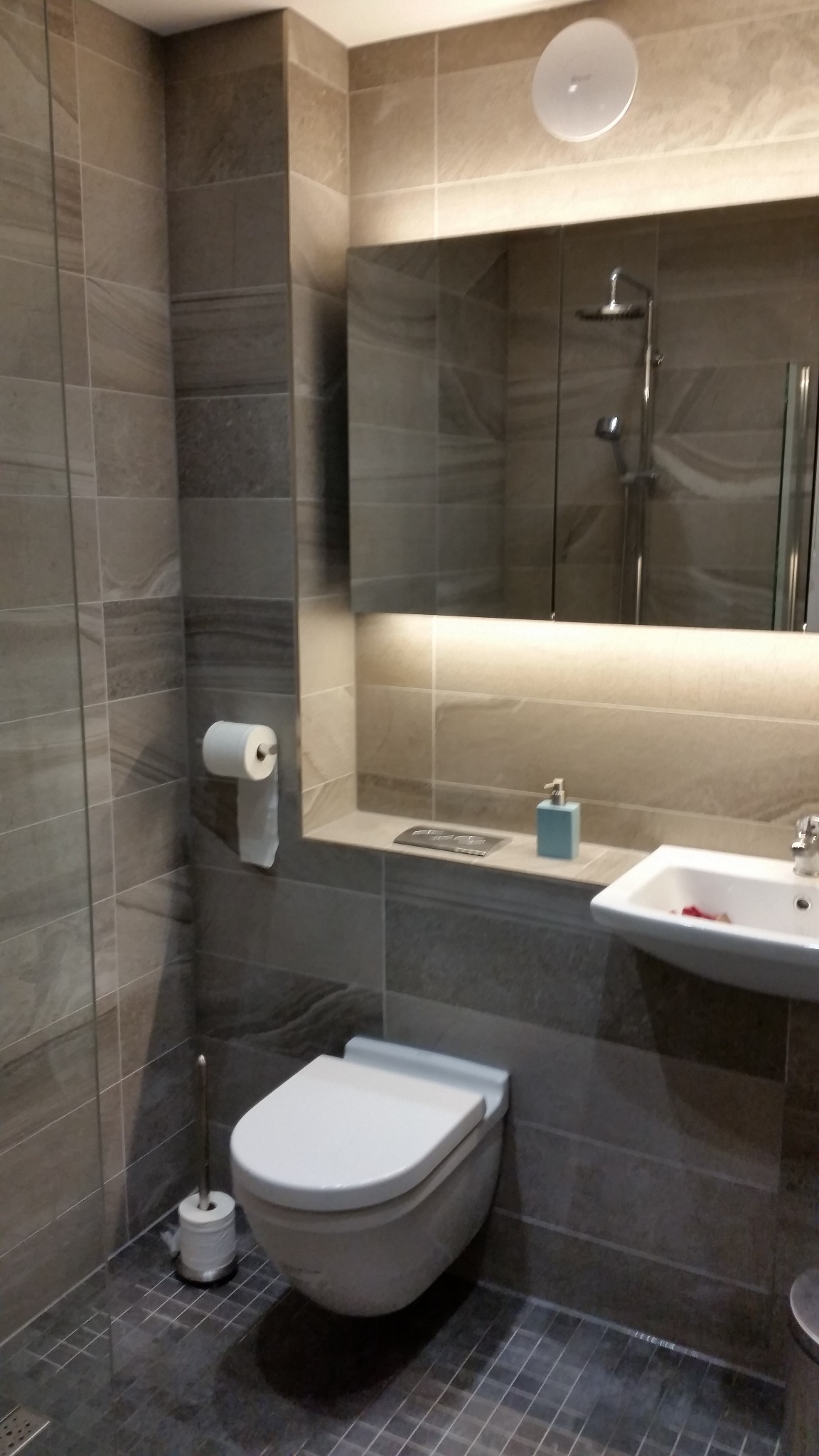 Nowoczesna łazienka w szarościach z podświetlanym lustrem, wiszącą toaletą i umywalką z dozownikiem na blacie.