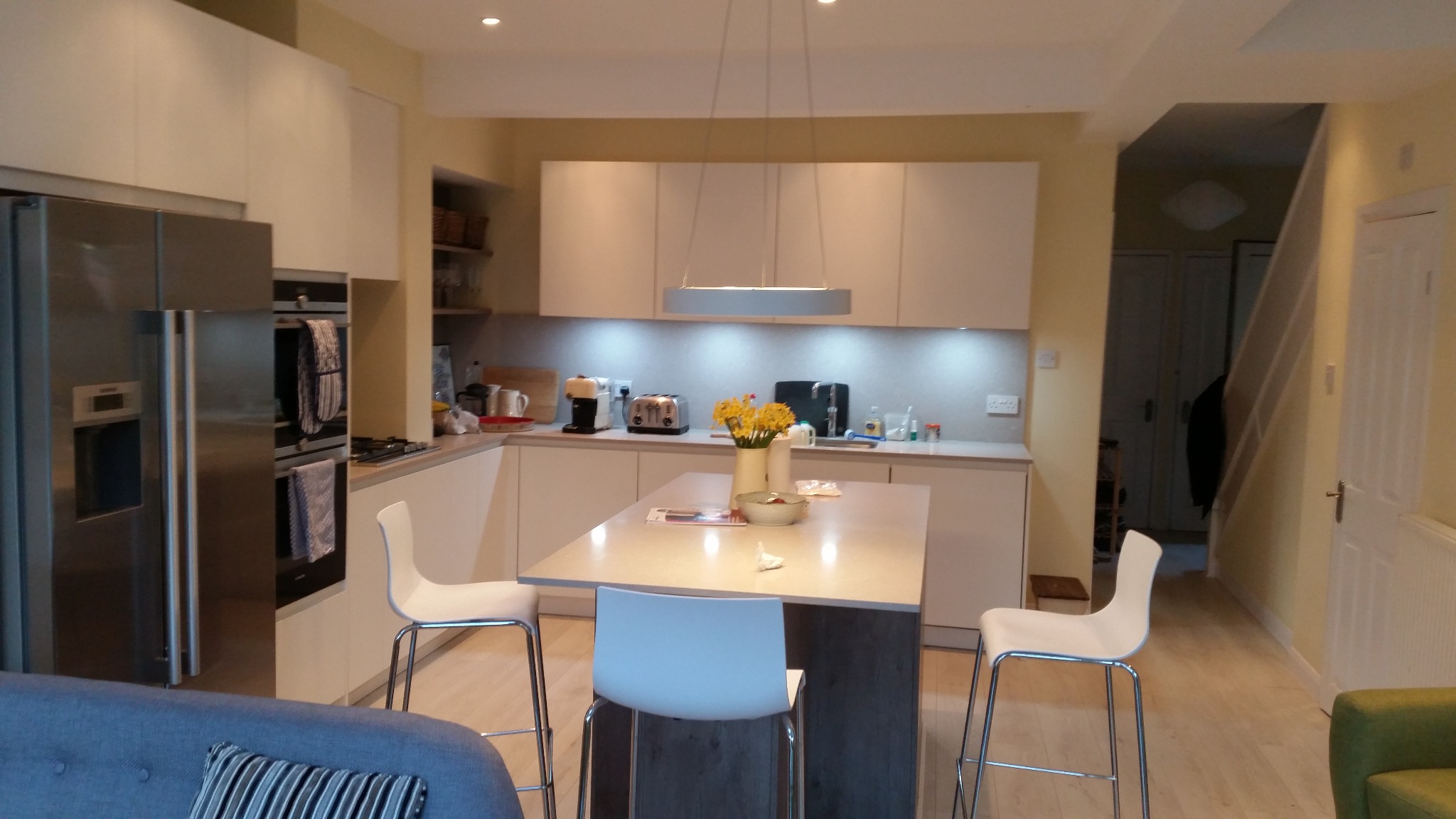 Jasna, nowoczesna kuchnia z wyspą, trzema białymi hokerami, stalową lodówką i oświetleniem LED pod szafkami, widok z salonu.