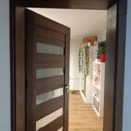 Ciemnobrązowe drzwi wewnętrzne z poziomymi przeszkleniami, częściowo otwarte, widok na pokój z panelami podłogowymi i białymi meblami.