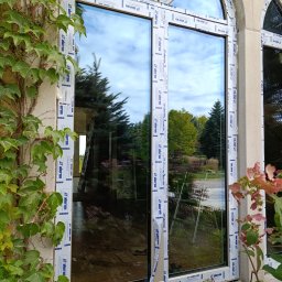 Adam - Nowo zamontowane okno PCV z folią ochronną producenta Aluron, widok z zewnątrz na ogród, telefon leżący na parapecie.