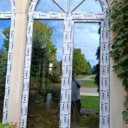 Adam - Nowe okno PCV z podziałem łukowym, zabezpieczone folią ochronną producenta, widok z zewnątrz, odbicie fotografa w szybie.