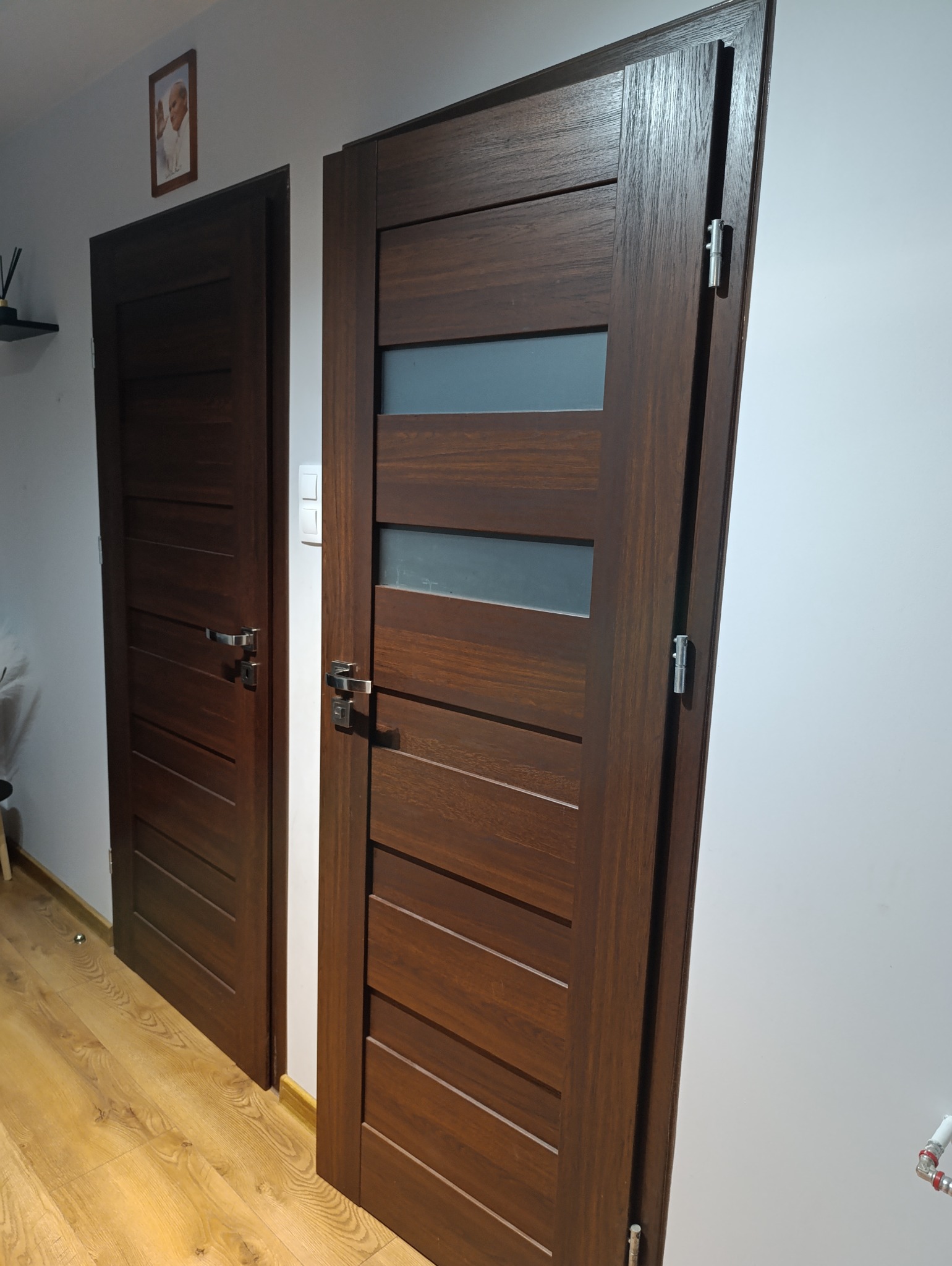 Dwa skrzydła drzwiowe w odcieniach brązu, jedno z przeszkleniami, zamontowane w jasnym wnętrzu z drewnianą podłogą; jedno skrzydło jest otwarte, ukazując zawiasy.