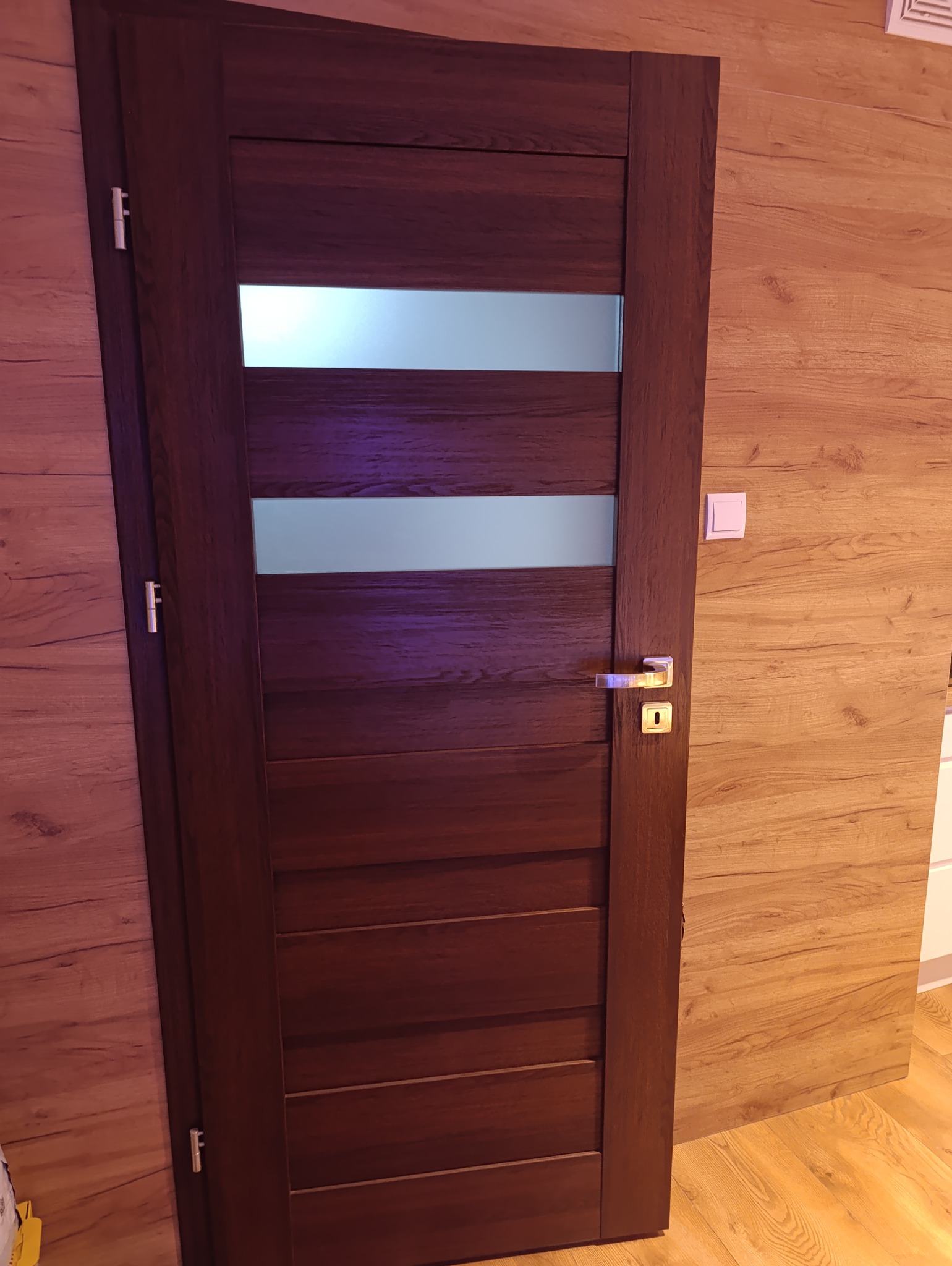 Ciemnobrązowe drzwi wewnętrzne z poziomymi panelami i przeszkleniami, z klamką w kolorze złotym, zamontowane w pomieszczeniu z panelami ściennymi o wzorze drewna.