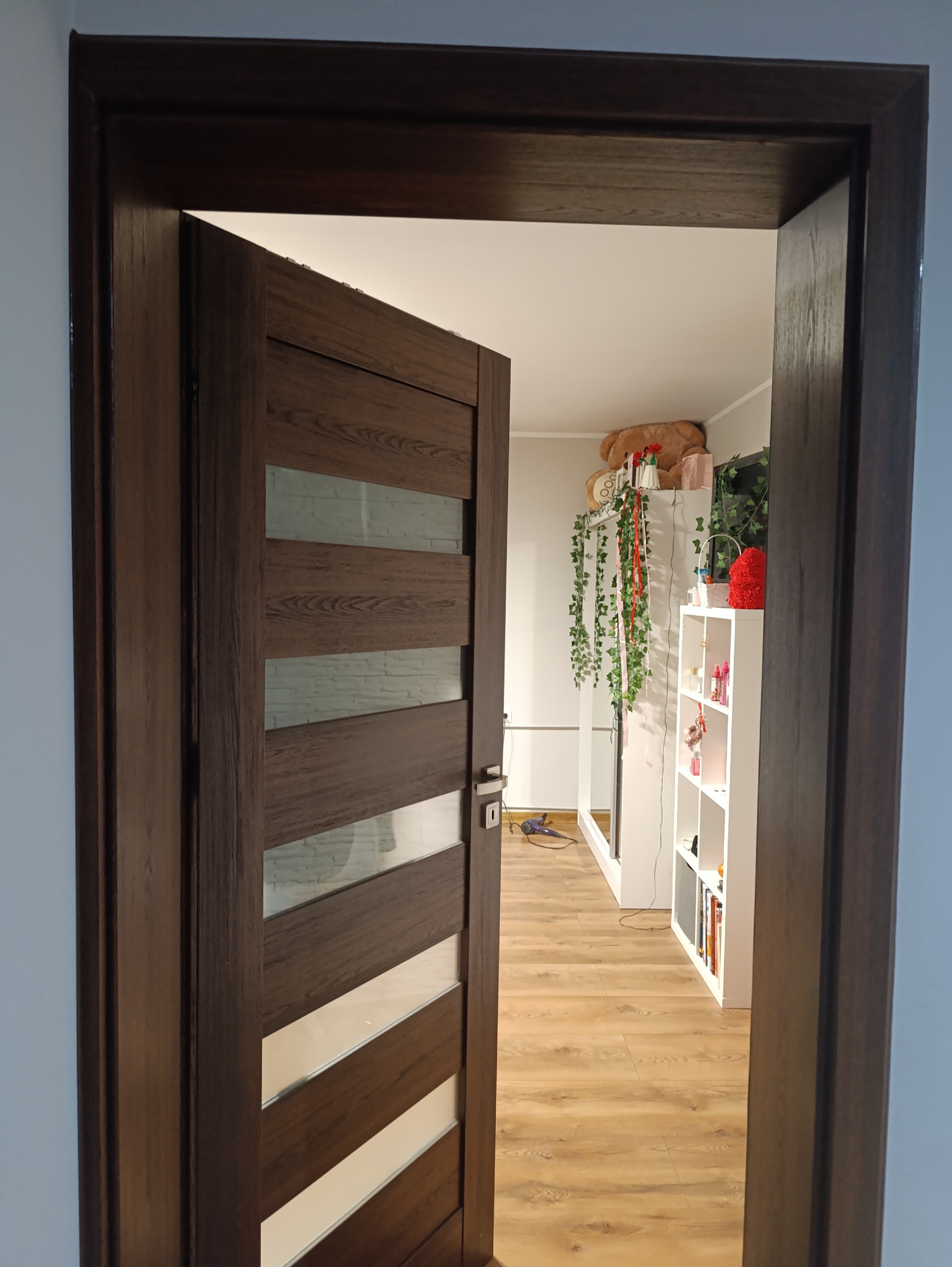 Ciemnobrązowe drzwi wewnętrzne z poziomymi przeszkleniami, częściowo otwarte, widok na pokój z panelami podłogowymi i białymi meblami.