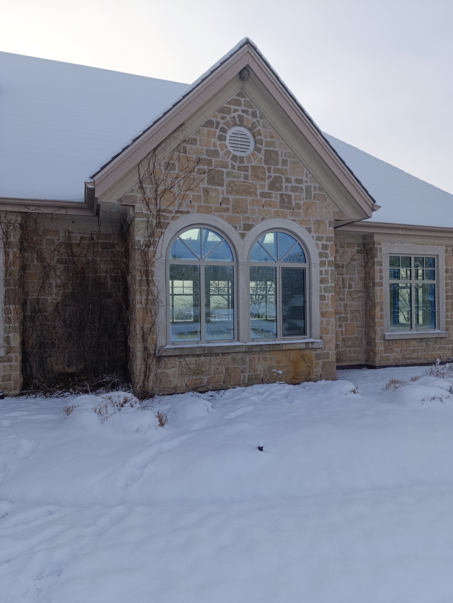 Kamienny dom z dwoma łukowymi oknami i jednym prostokątnym, pokryty śniegiem, z widocznymi śladami na śniegu.