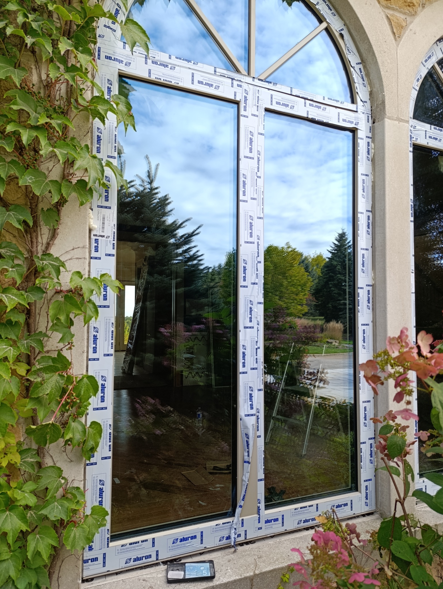 Nowo zamontowane okno PCV z folią ochronną producenta Aluron, widok z zewnątrz na ogród, telefon leżący na parapecie.