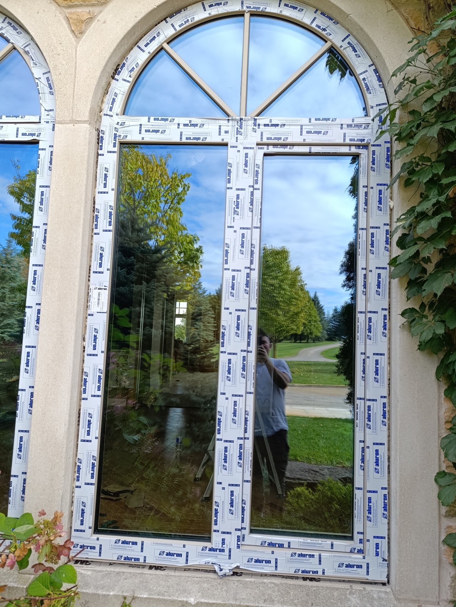 Nowe okno PCV z podziałem łukowym, zabezpieczone folią ochronną producenta, widok z zewnątrz, odbicie fotografa w szybie.