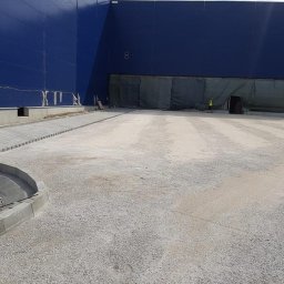 Dariusz Kisielewski Przedsiębiorstwo Budowlano-Usługowe DAR-BRUK - Przygotowanie terenu pod parking: utwardzona nawierzchnia z podsypką, fragment ułożonej kostki przy budynku w tle, pracownik w odblaskowej kamizelce w oddali.