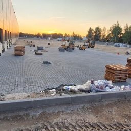 Dariusz Kisielewski Przedsiębiorstwo Budowlano-Usługowe DAR-BRUK - Trwa układanie szarej kostki brukowej na dużej powierzchni w Oświęcimiu, widoczne palety z kostką, pracownik w czerwonym kasku i maszyny budowlane w tle, zachodzące słońce.