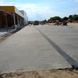 Dariusz Kisielewski Przedsiębiorstwo Budowlano-Usługowe DAR-BRUK - Nowo ułożona, szara kostka brukowa przed budynkiem handlowym z widocznymi maszynami budowlanymi w tle.