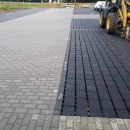 Widok z góry na częściowo ułożoną nawierzchnię parkingu z kostki brukowej w odcieniach szarości i czerni, z żółtą ładowarką teleskopową w tle i samochodami na dalszym planie.