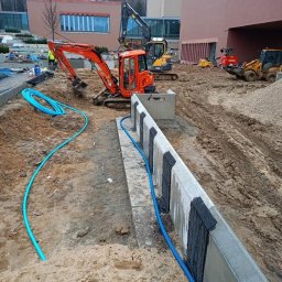 Dariusz Kisielewski Przedsiębiorstwo Budowlano-Usługowe DAR-BRUK - Budowa systemu nawadniania: pomarańczowa mini koparka w wykopie obok betonowego murku z zabezpieczeniem hydroizolacyjnym, niebieskie rury nawadniające ułożone wzdłuż wykopu, w tle inne maszyny...