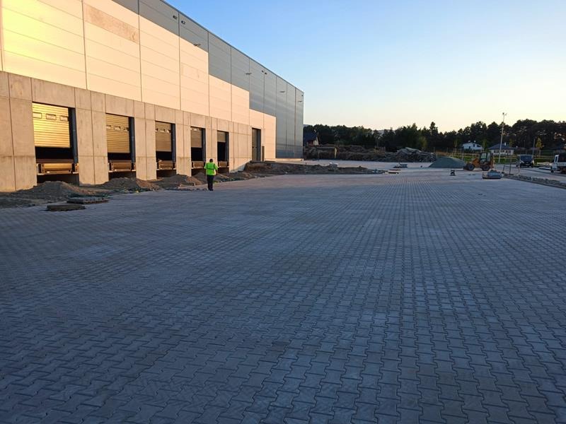 Rozległy plac wyłożony szarą kostką brukową typu 'psi kość' przed budynkiem magazynowym z otwartymi bramami załadunkowymi; w tle widoczny pracownik w kamizelce odblaskowej i sprzęt budowlany.