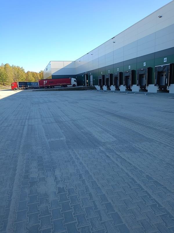 Rozległy plac wyłożony szarą kostką brukową przed budynkiem magazynowym z rampami załadunkowymi i zaparkowanymi ciężarówkami w tle.