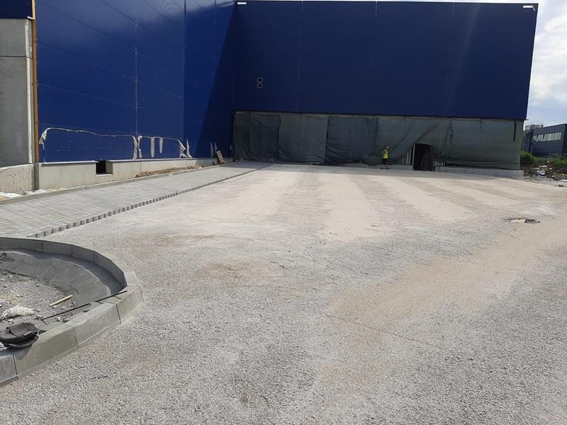 Przygotowanie terenu pod parking: utwardzona nawierzchnia z podsypką, fragment ułożonej kostki przy budynku w tle, pracownik w odblaskowej kamizelce w oddali.