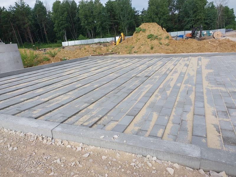 Szara, prostokątna kostka ułożona w rzędach, z piaskiem między szczelinami, obrzeża betonowe, w tle kopiec ziemi, maszyny budowlane i drzewa.