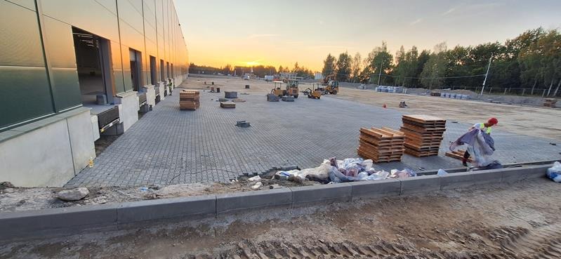 Trwa układanie szarej kostki brukowej na dużej powierzchni w Oświęcimiu, widoczne palety z kostką, pracownik w czerwonym kasku i maszyny budowlane w tle, zachodzące słońce.