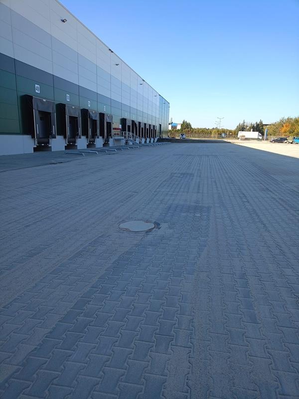 Rozległy plac wyłożony szarą kostką brukową typu 'I', przylegający do hali magazynowej z rampami załadunkowymi w słoneczny dzień.
