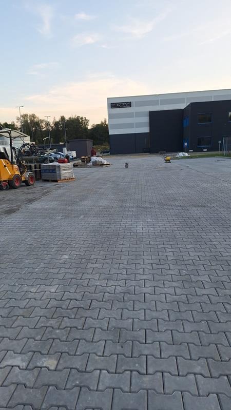 Ułożona szara kostka brukowa na parkingu przed budynkiem magazynowym, z widocznym sprzętem budowlanym i stertą kostki na paletach w tle, wczesnym wieczorem.