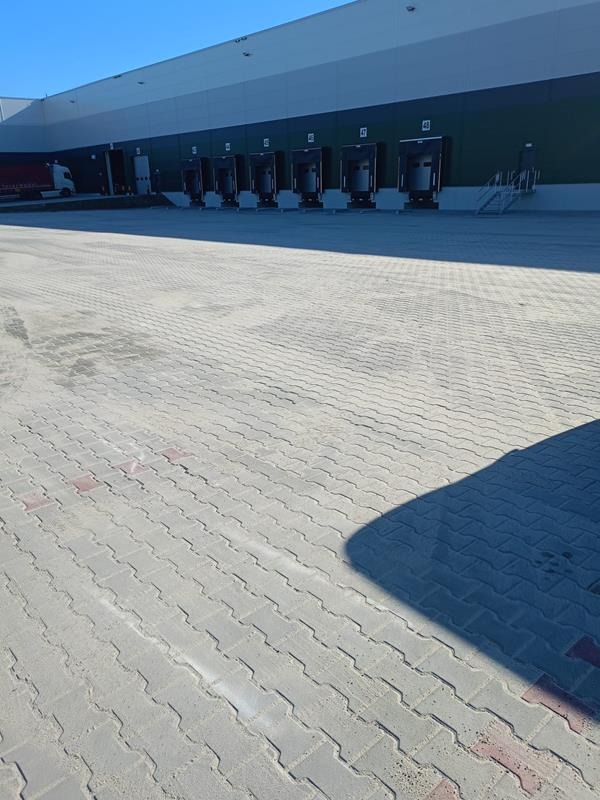 Szara nawierzchnia z kostki brukowej przed budynkiem magazynowym z rampami załadunkowymi, widoczny cień.