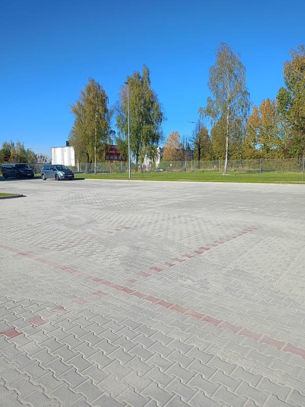 Rozległy parking wyłożony szarą kostką z czerwonymi liniami wyznaczającymi miejsca postojowe, samochody w tle, błękitne niebo i drzewa z jesiennymi liśćmi.