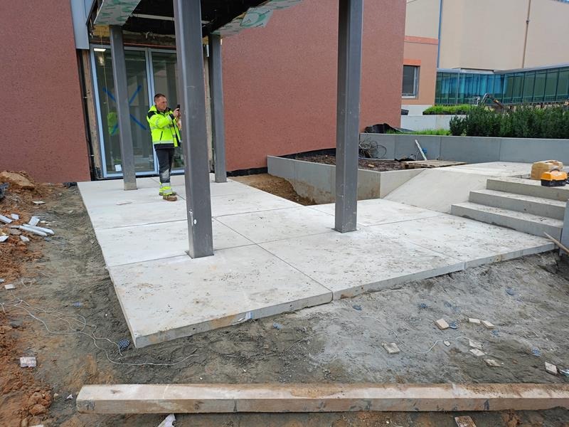 Ułożony chodnik z betonowych płyt w przestrzeni publicznej, pracownik w odblaskowej kurtce robi zdjęcie telefonem, widoczne przygotowania do dalszych prac ziemnych.