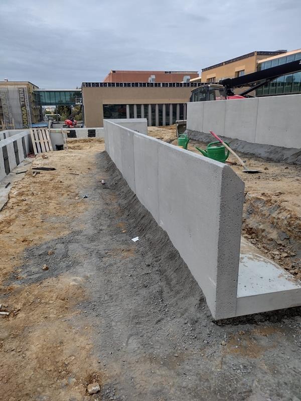Wznoszenie betonowego murku oporowego na placu budowy, widoczne dwie zielone konewki i łopata, w tle budynek z przeszklonym łącznikiem.