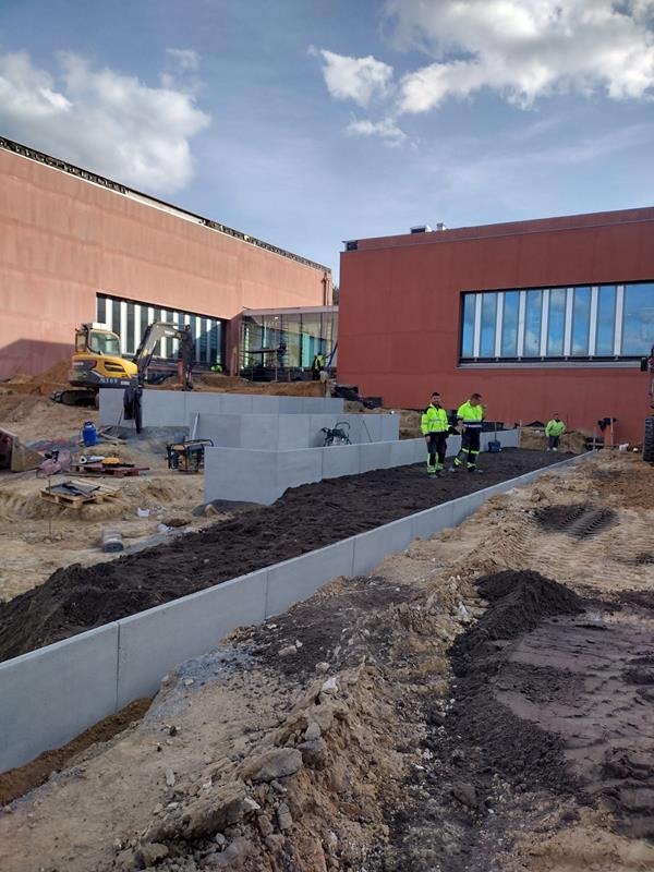 Budowa: Wykop z betonowym obrzeżem, widoczny żółty mini-koparka i pracownicy w odblaskowych kurtkach na tle budynku z czerwonej cegły pod błękitnym niebem.