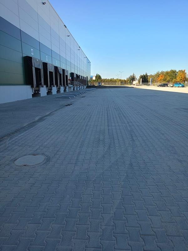 Rozległy plac wyłożony szarą kostką brukową typu I, przylegający do hali magazynowej z rampami załadunkowymi, widok z niskiej perspektywy w słoneczny dzień.