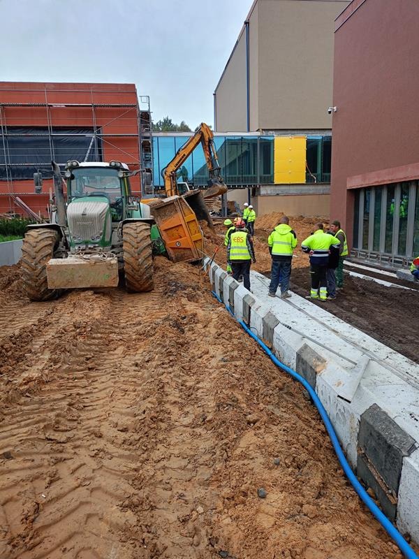 Tractor i koparka podczas prac ziemnych na budowie, widoczni pracownicy w odblaskowych kamizelkach. Świeżo wykopany rów z ułożonymi prefabrykatami betonowymi i niebieską rurą.