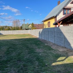 Patryk Wiśniewski - Długi, szary, betonowy płot z fakturą kamienia oddziela posesję z trawnikiem, w tle domy i błękitne niebo z chmurami. Widok z perspektywy.