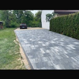 Patryk Wiśniewski - Ułożona szara kostka brukowa tworząca podjazd do domu z zielonym żywopłotem. W tle zaparkowany samochód i fragment drewnianej konstrukcji.