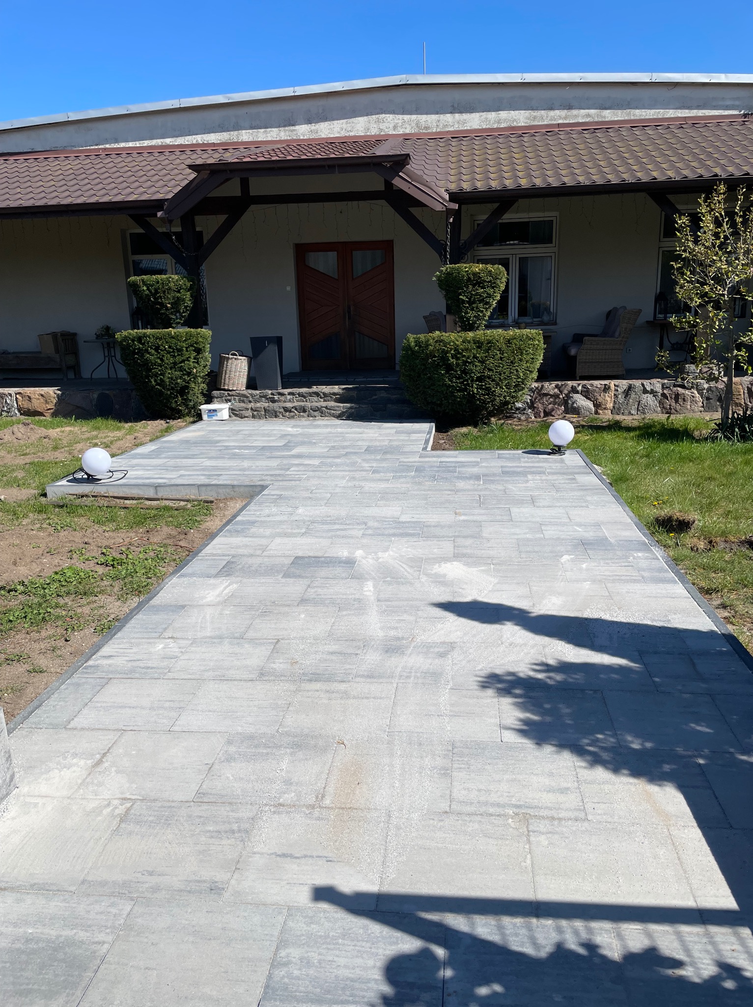 Ułożona ścieżka z szarych płyt brukowych prowadząca do domu z drewnianym gankiem, oświetlona kulistymi lampami. Widok na elewację budynku i zadbany trawnik.