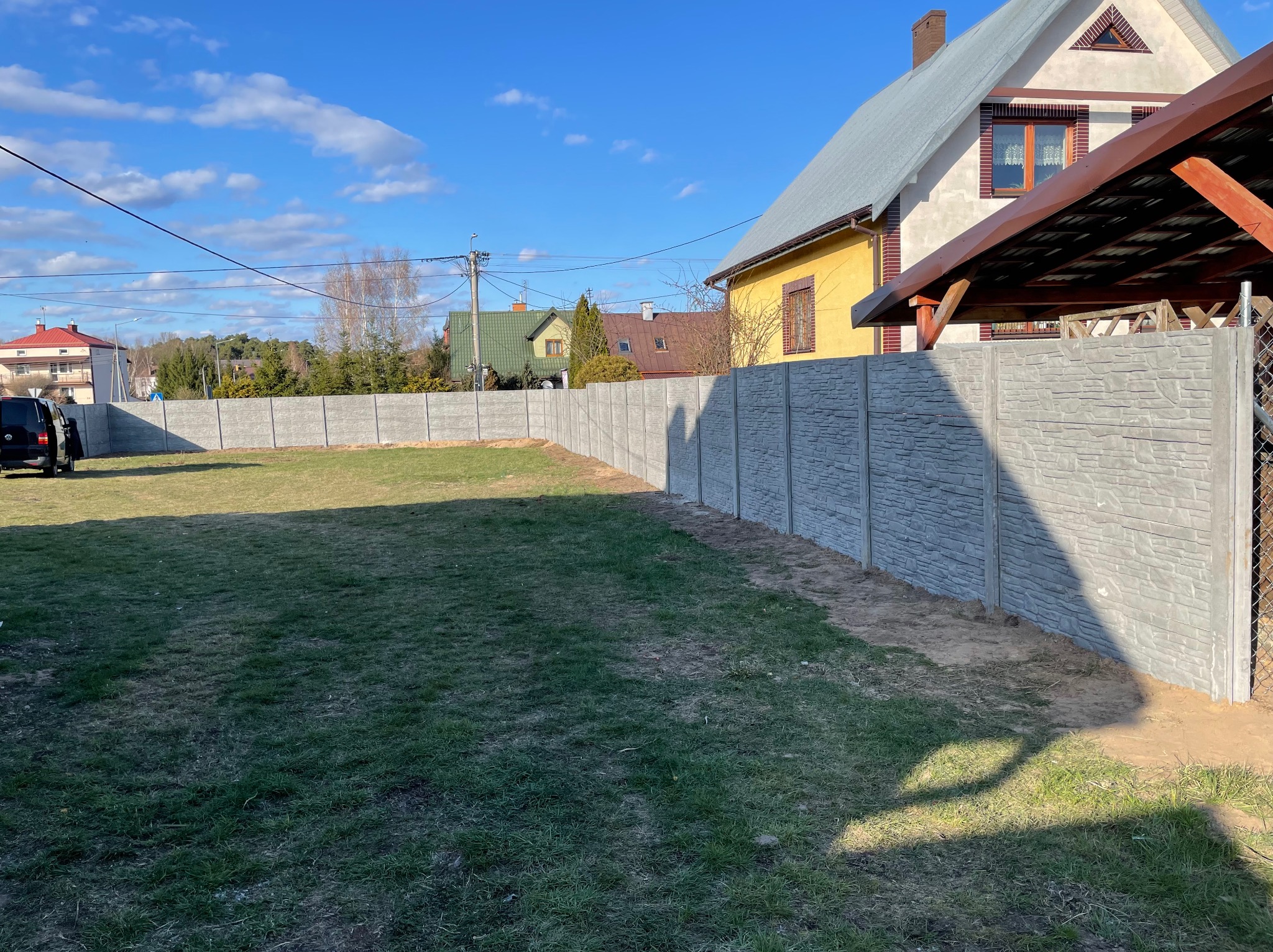 Długi, szary, betonowy płot z fakturą kamienia oddziela posesję z trawnikiem, w tle domy i błękitne niebo z chmurami. Widok z perspektywy.