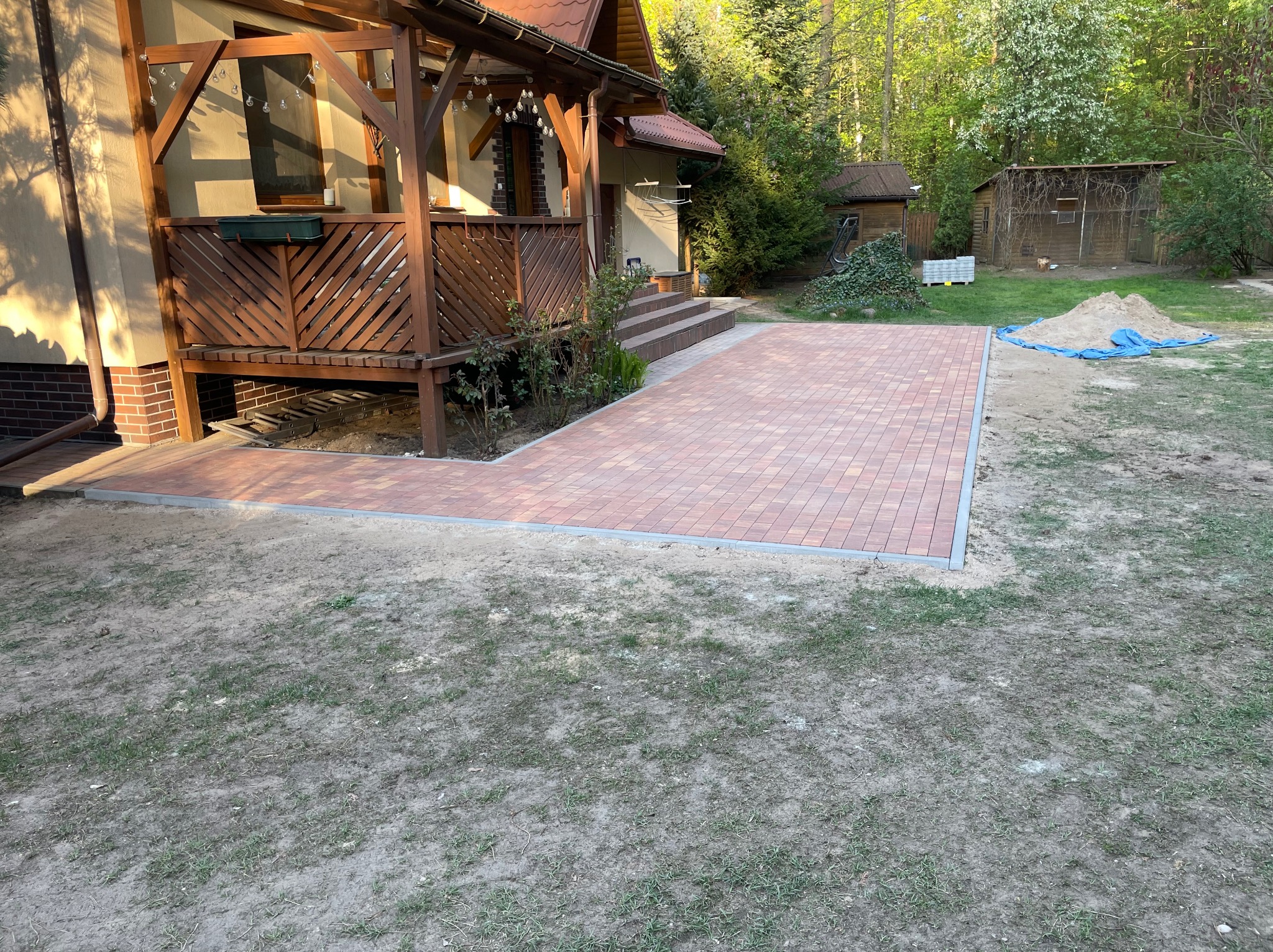 Nowo ułożona, czerwono-brązowa kostka brukowa prowadząca od drewnianego tarasu domu w kierunku trawnika, z widoczną stertą piasku w tle.