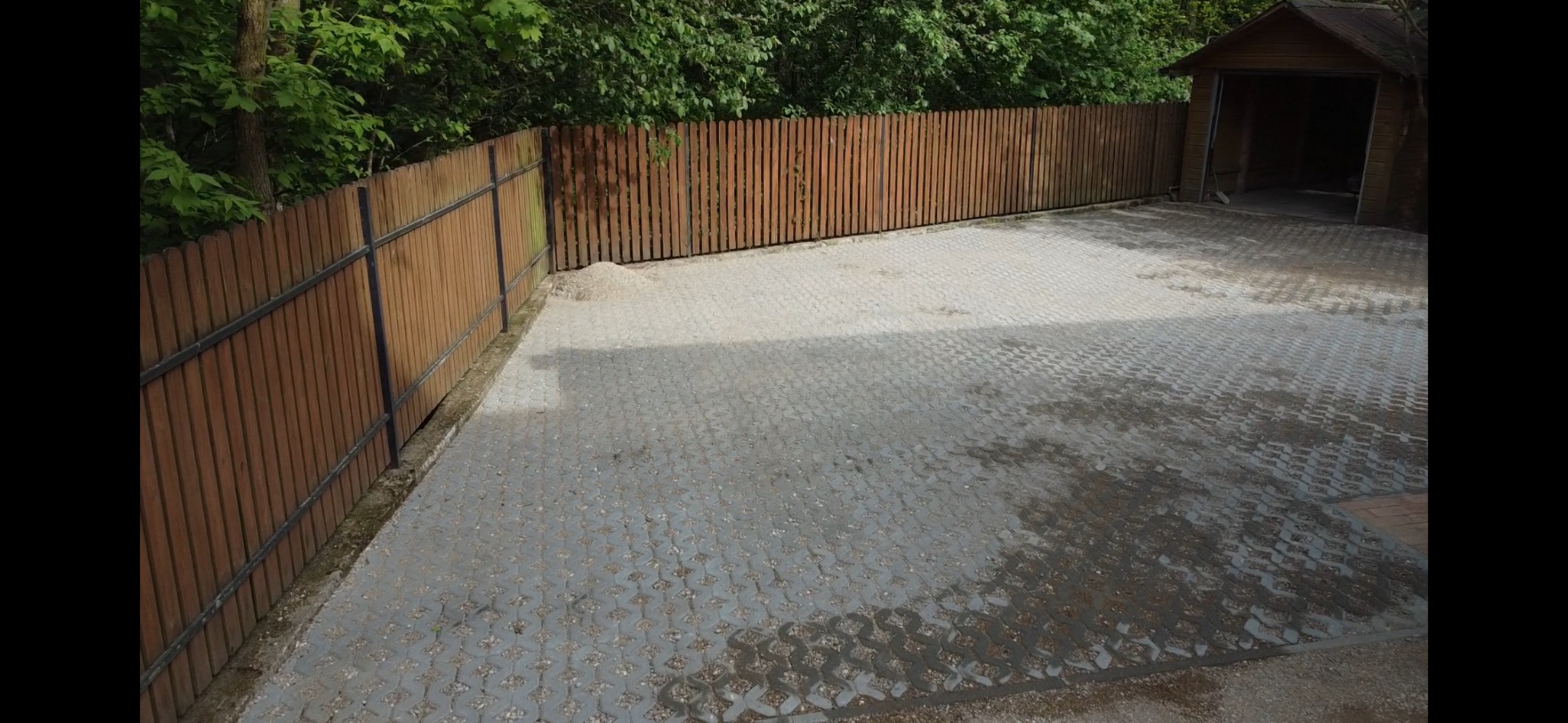 Ułożona szara kostka brukowa typu ażur, obramowana drewnianym ogrodzeniem z metalową ramą. W tle widoczny garaż i zieleń drzew.