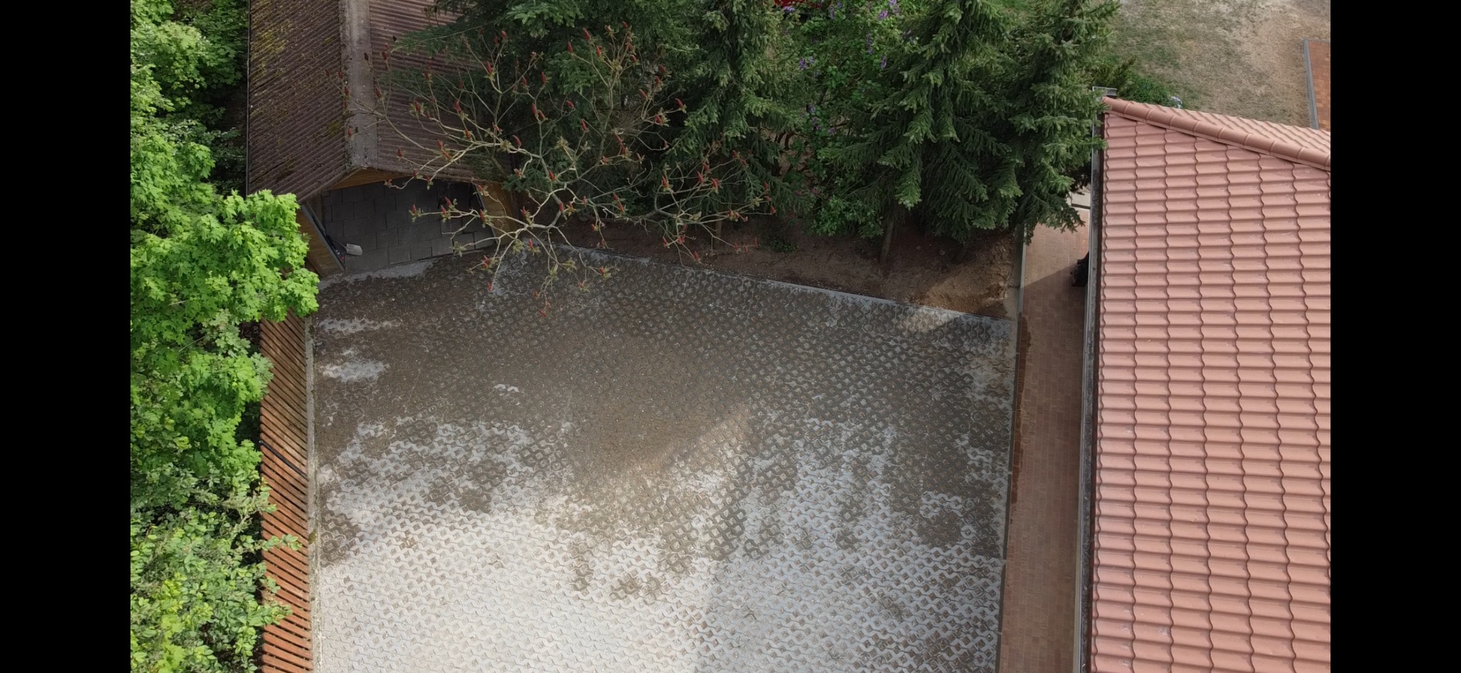 Widok z góry na ułożoną kostkę brukową na podjeździe, częściowo mokrą, otoczoną zielenią i budynkami z czerwoną dachówką. Kąt ujęcia podkreśla wzór kostki.