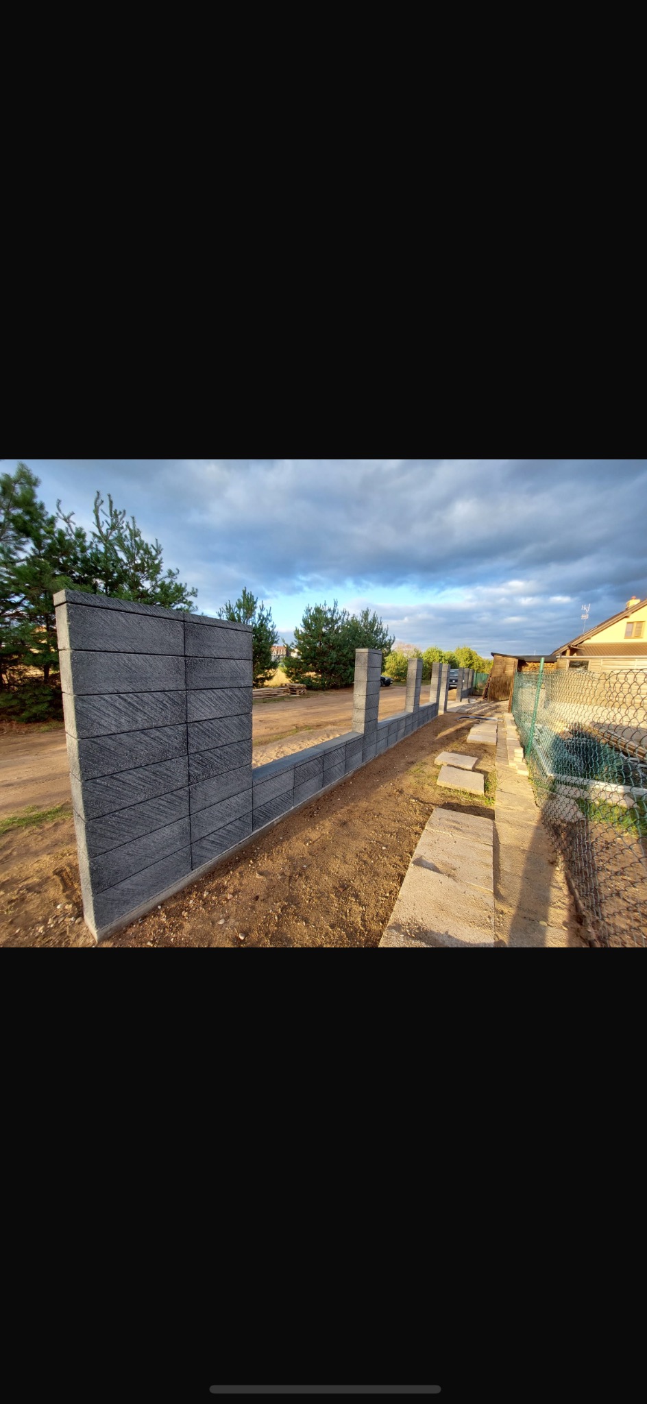 Wznoszenie muru z szarej kostki betonowej na posesji, widok z boku. Fragment gotowego ogrodzenia z filarami i podmurówką w trakcie budowy, z widocznym terenem i zielenią w tle.