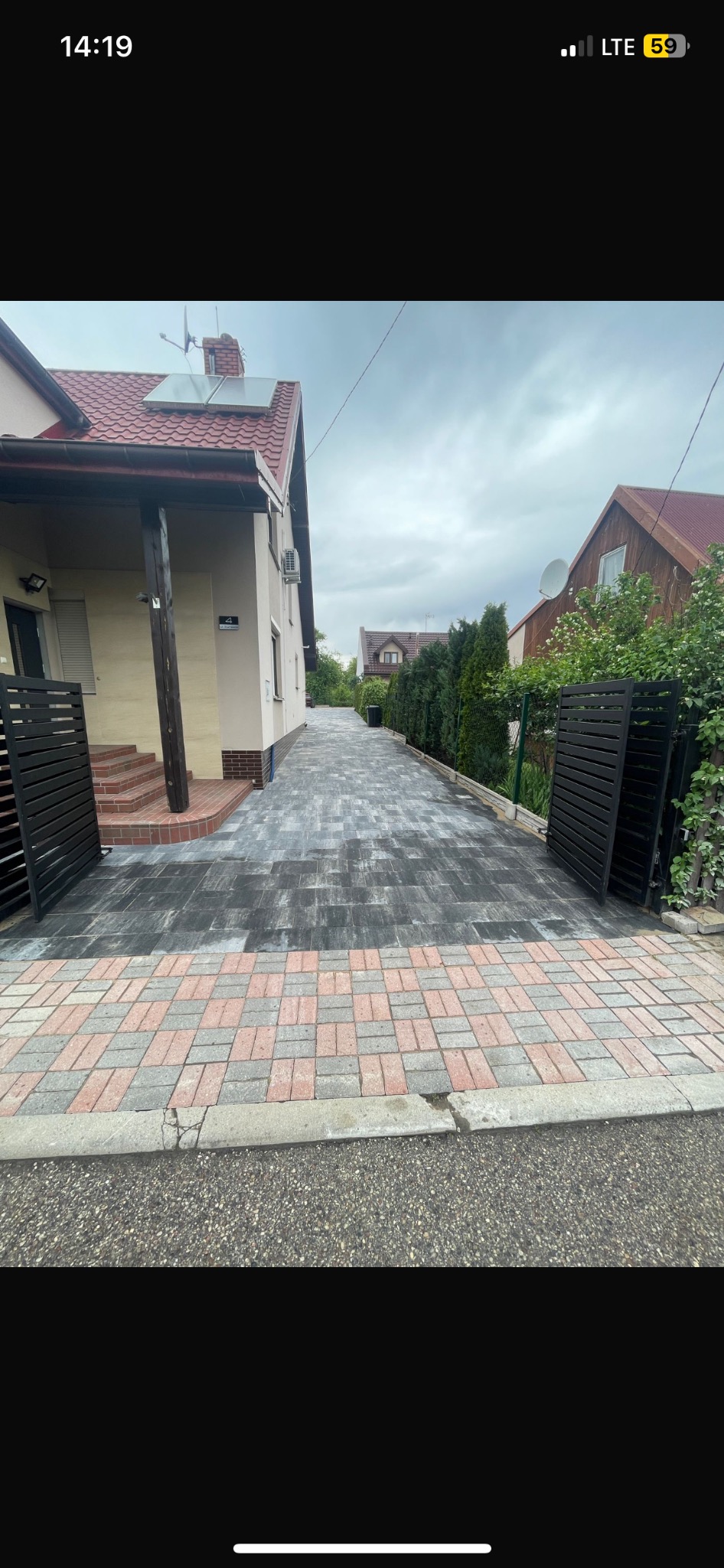 Ułożona kostka brukowa w odcieniach szarości i czerwieni tworzy wzór na podjeździe. Nowoczesne, czarne ogrodzenie panelowe po bokach. Dom z panelami słonecznymi w tle.