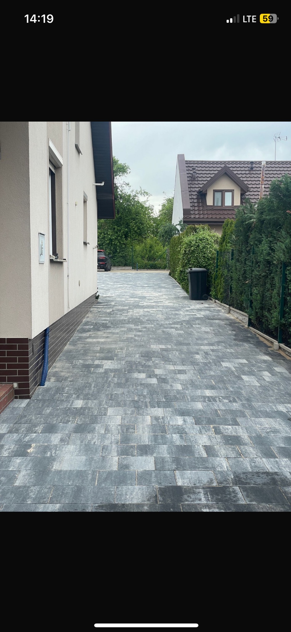 Długa, szara ścieżka z kostki brukowej prowadząca wzdłuż domu z brązową podmurówką i zielonego żywopłotu, aż do drugiego domu w tle.